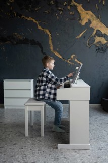 A young child sits at a sleek white piano, focused on playing music. The modern interior features a