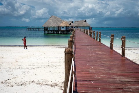 A long wooden pier leads to traditional thatched-roof bungalows over turquoise waters on a white