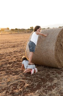 Two children enjoy a sunny day in a rural field. One climbs a large hay bale while the other