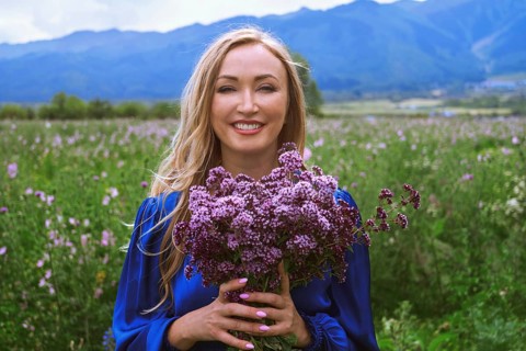 A joyful blond woman stands in a grassy field, smiling brightly while holding a bouquet of purple