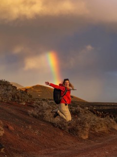 A woman in a red jacket jumps joyfully against a backdrop of a vivid rainbow in Timanfaya National