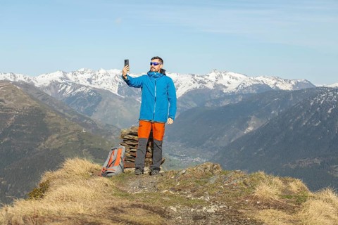 A man wearing a blue jacket and orange pants is trekking in the Pyrenees. He stands on a grassy