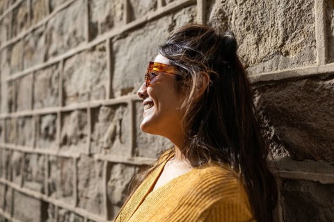 A Latin woman wearing sunglasses and a yellow sweater smiles while leaning against a textured stone