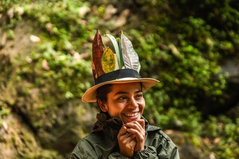 A young, joyful Ecuadorian woman wearing a hat adorned with tropical leaves smiles during an
