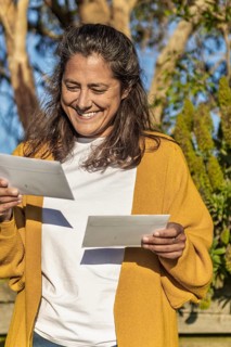 A woman stands in a sunlit Australian garden, smiling while holding two letters. Her yellow sweater