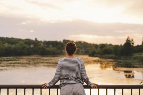 A woman stands at a railing, gazing towards a serene lake during sunset. The scene evokes a sense