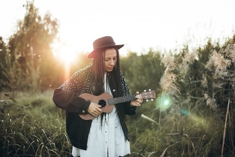 A woman with braided hair and a hat plays a ukulele amidst a serene field at sunset, surrounded by