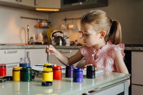 A young girl, dressed in pink, paints with bright colors at a kitchen table. Paint jars are