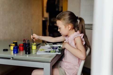 Young girl intently paints with vibrant colors at a home art table, surrounded by paint jars. She