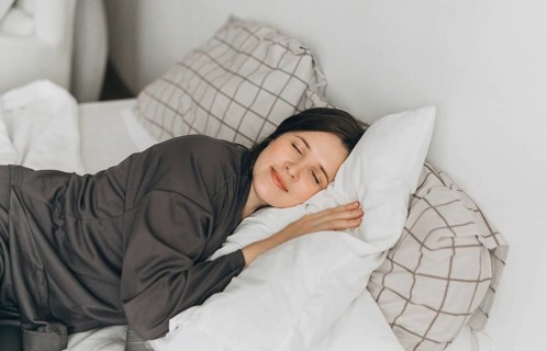 A young woman in comfortable loungewear lies on a white bed, resting her head on soft pillows with