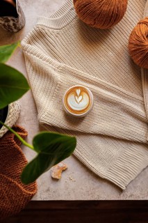 Flat lay of a warm latte on a knitted sweater surrounded by brown yarn balls and green leaves. The
