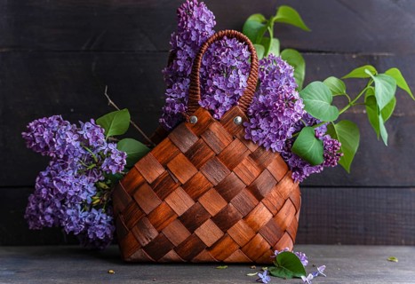 A rustic woven basket filled with vibrant purple lilacs against a dark wooden background. The lush