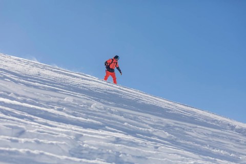 A snowboarder dressed in bright orange gear glides down a snowy slope against a clear blue sky. The