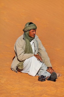 A man sits relaxed on the sand in the desert wearing traditional clothing and a green headscarf, A