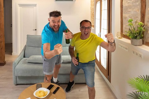 Two male friends celebrating a goal with excitement while watching a sports match at home. Raised