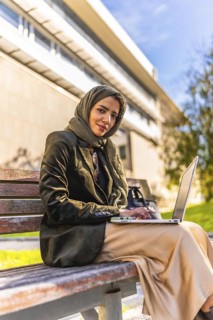 Vertical photo of a young Arab woman in hijab working using laptop sitting on park bench