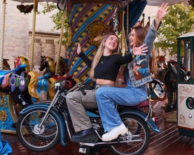 Two smiling friends enjoy a playful moment on a carousel motorcycle in an amusement park,