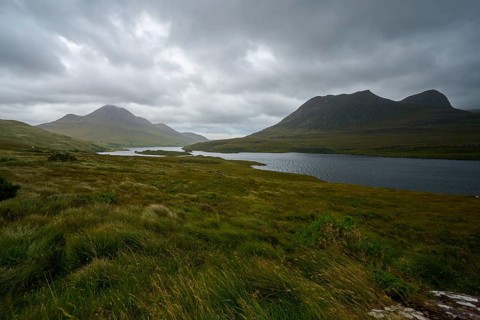 Wide grassland in front of a lake and mountains under a dark, thick sky, Loch Bad a Ghaill,