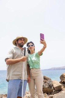 A blind man with a cane and a woman stand close together, capturing a selfie by the coast. The