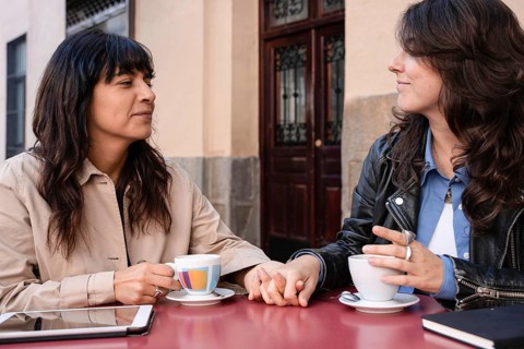 A multiethnic lesbian couple holds hands and shares a coffee at an outdoor cafe. Their interaction
