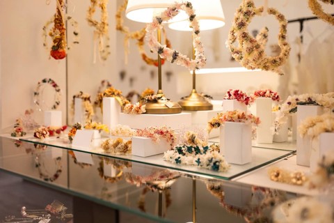 A variety of handcrafted floral headpieces are elegantly displayed on glass shelves. Soft lighting