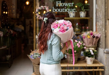 A woman stands in a flower shop, holding a vibrant bouquet of pink and white flowers. The shop is