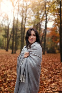 A woman wrapped in a cozy gray blanket smiles gently amidst a forest filled with colorful autumn