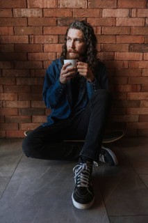 A man with long hair and a beard sits on the floor against a brick wall, holding a cup of coffee.
