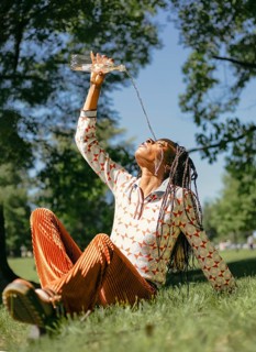 African American woman with braided hair enjoys water in nature, sitting on grass in a sunlit park.