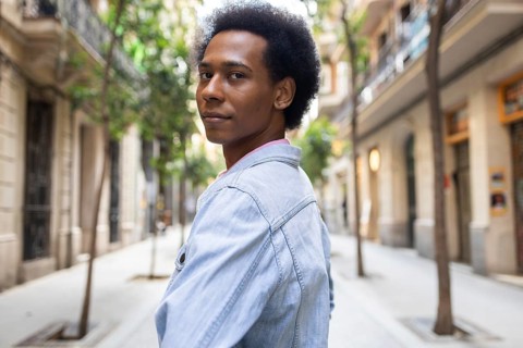 A non-binary person of African American descent walks confidently down a sunlit urban street,