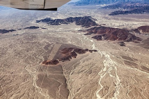 Bird's-eye view of desert landscape with mountain ranges and part of an airplane, the geoglyphs and