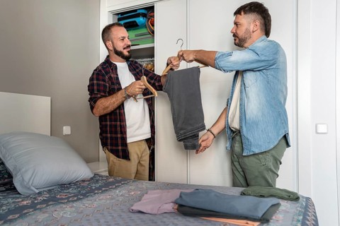 A gay couple stands in their bedroom, selecting clothes from a wardrobe. One holds a pair of pants