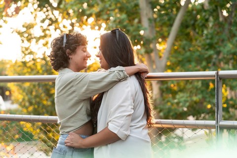 A lesbian couple shares a romantic embrace in a sunlit park. They look into each other's eyes,