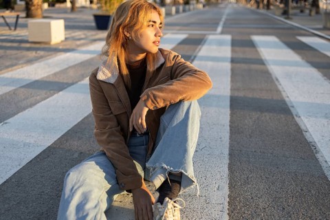 A Mixed-race woman wearing a brown jacket and jeans sits casually on a crosswalk, gazing into the
