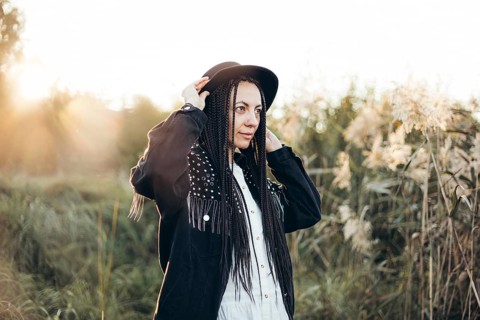 A woman in a stylish outfit and hat stands amidst tall grasses at sunset. The sun casts a warm