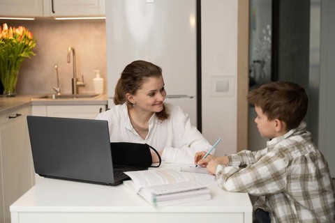A mother guides her son in a home learning setup, featuring a laptop and books on a kitchen table.