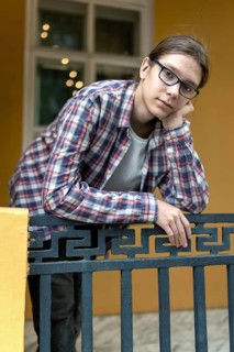 A teenage boy with long hair rests his face on his hand while leaning on a decorative railing. He