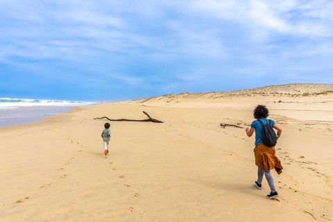 Mother and son running and playing on a wide sand beach near the ocean waves, with sand dunes in