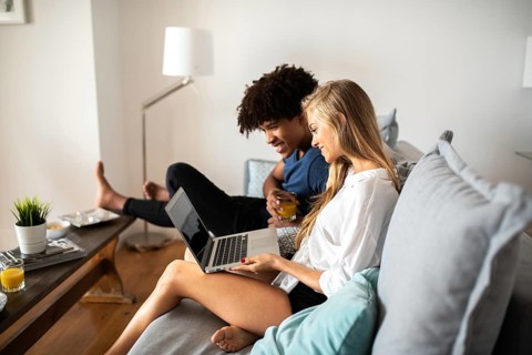 A couple sits comfortably on a couch in their living room, sharing a laptop. Natural light enhances