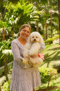 Smiling mature woman holding her small white poodle dog wearing a pink dress, enjoying a sunny day