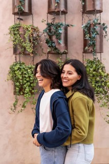 A cheerful lesbian couple embraces, smiling brightly amidst a backdrop of greenery and hanging