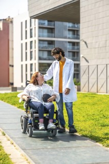 Doctor assisting a female patient in an electric wheelchair, strolling together outdoors in a park,