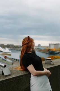 A red-haired woman in a black top and white skirt leans on a stone wall, enjoying the scenic view