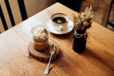 A cozy wooden table displays a jar dessert, a cup of coffee on a saucer, and a small rustic vase