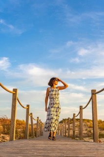 Woman in a summer dress and sunglasses walking along a wooden boardwalk through sand dunes,
