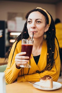 A woman in a yellow sweater and dreadlocks enjoys a drink through a straw, sitting at a table with