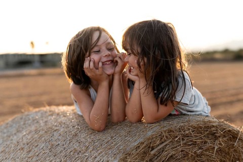 Two young children share a joyful moment on a bale of hay in a sunny rural field. Their laughter
