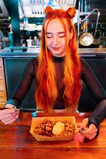 A woman with long, vibrant hair savors a dish with chopsticks at an Asian food market She smiles,