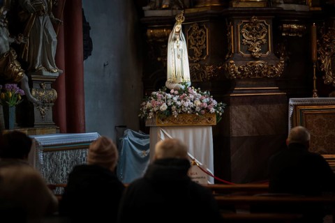Devout people praying in a dim church before a flower-decorated statue of the Virgin Mary during a