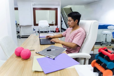 A dedicated physiotherapist in a modern Ecuadorian clinic works on a laptop. The setting includes